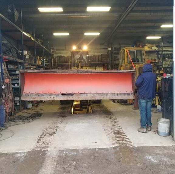 East-West snowplow repair shop in Toronto with a plow blade on the bench and a mechanic working